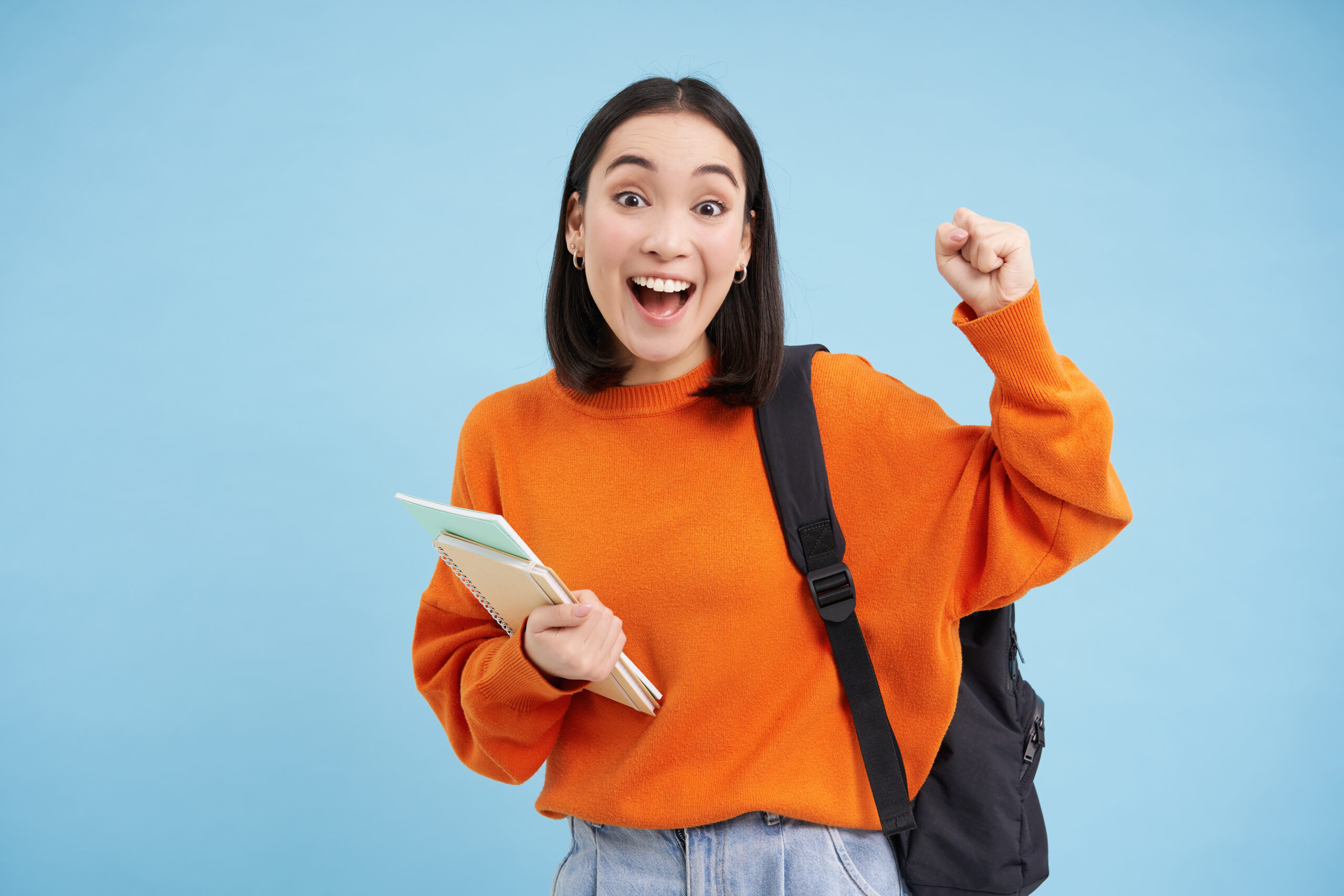 Enthusiastic girl student, celebrating, wearing backpack and holding notebooks, college homework, standing over blue background